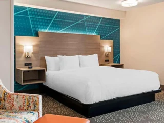 Modern hotel room with a large bed, wooden headboard, and abstract blue wall design. Two wall-mounted lamps are on either side of the bed. Patterned armchair and orange ottoman in foreground.