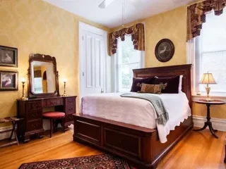 A bedroom with a wooden bed, white bedding, and decorative pillows. A matching dresser with a mirror and a cushioned stool are on the left. Two windows with drapes are in the room.