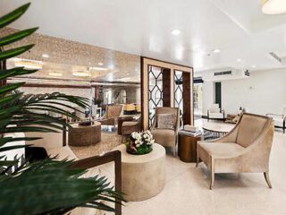 Modern lobby with beige armchairs, a round table, and decorative plants. Large mirrors and elegant lighting enhance the spacious and welcoming atmosphere.