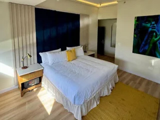 A neatly made bed with white linens and a yellow accent pillow in a well-lit bedroom with modern decor, flanked by a small wooden side table and abstract wall art.