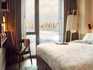 A cozy hotel room with a bed, chair, and clothes rack by a window showing a city skyline view.