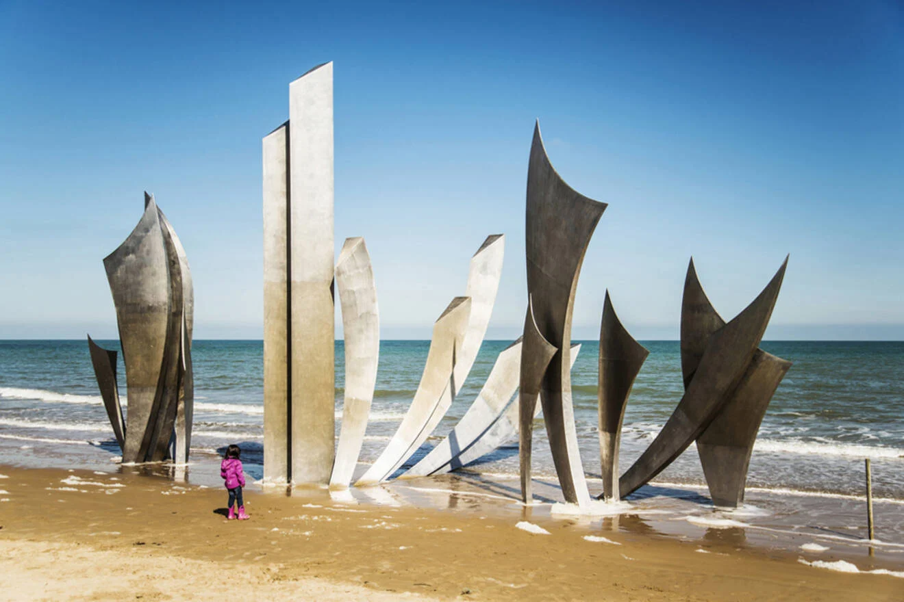 A child in a pink jacket stands on a sandy beach, looking at a modern metal sculpture with tall, curved panels near the ocean under a clear blue sky in one of the Normandy beaches