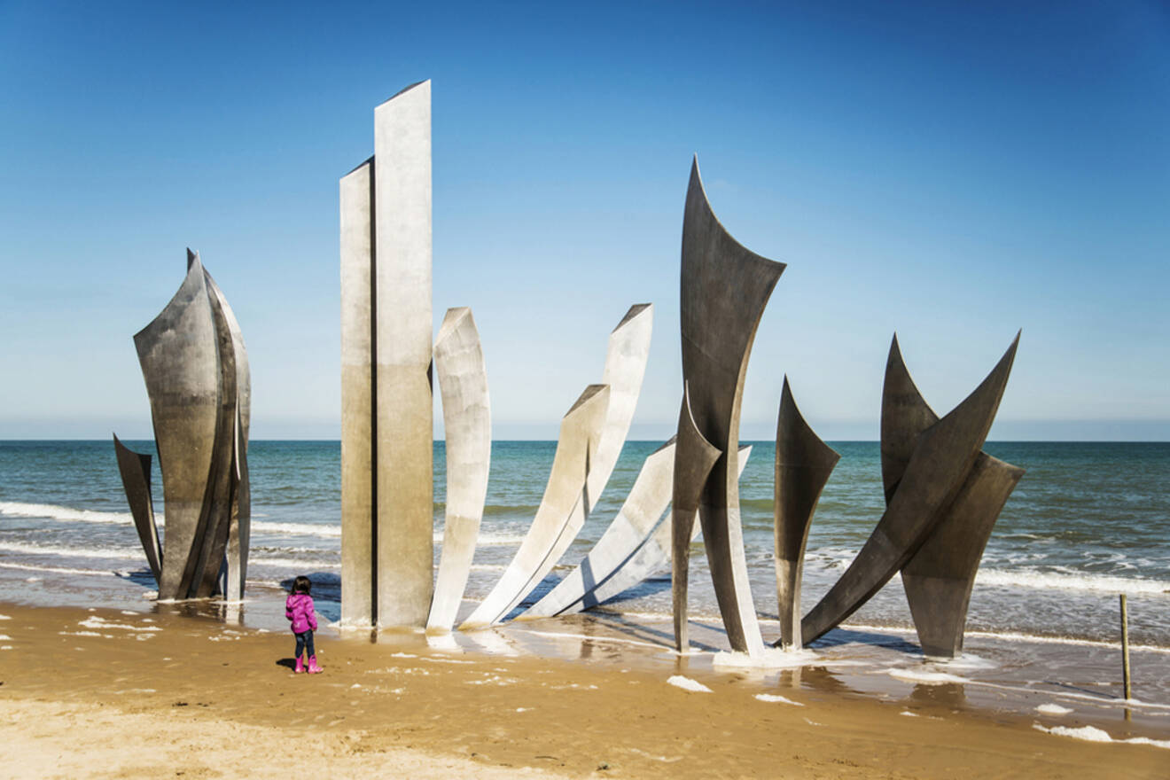 A child in a pink jacket stands on a sandy beach, looking at a modern metal sculpture with tall, curved panels near the ocean under a clear blue sky in one of the Normandy beaches 