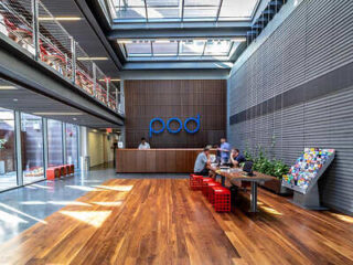 A modern office lobby with wooden flooring, a front desk, "pod" logo on the wall, people seated at a table, and a magazine rack to the side. Large windows provide natural light.