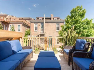 Rooftop deck with blue-cushioned wicker furniture, overlooking a brick building and greenery under a clear sky.