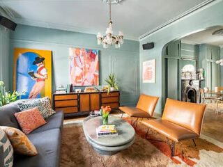 A colorful living room with modern decor, featuring a gray sofa, two tan chairs, a marble oval coffee table, and vibrant artwork on green walls.