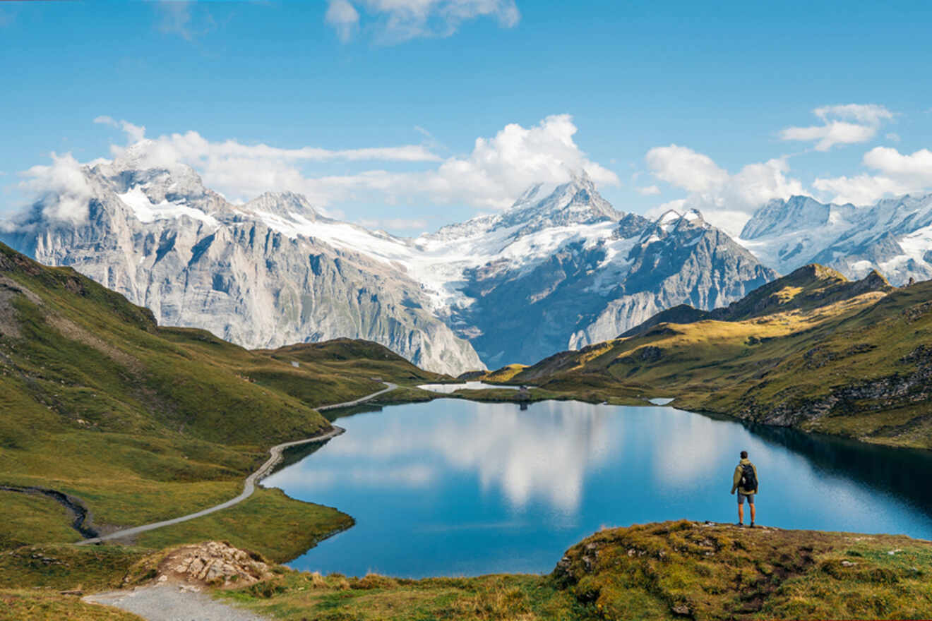 A hiker stands by a calm mountain lake, surrounded by greenery and snow-capped peaks under a partly cloudy sky.