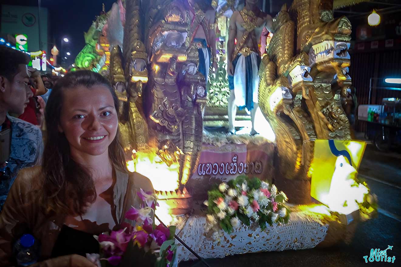 The writer of the post holding flowers stands in front of ornate, illuminated parade float with golden details at night in Chiang-Mai