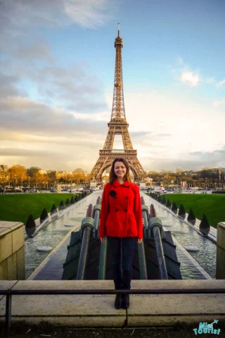 The writer of the post in a red coat stands in front of the Eiffel Tower with fountains in the foreground.