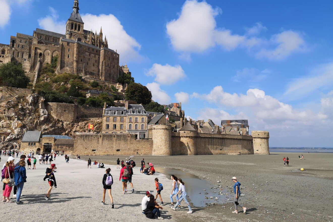 Tourists explore the sandy area leading to Mont Saint-Michel, a historic island with a medieval abbey and stone buildings, under a partly cloudy sky.