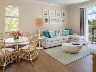 A bright living room with a gray sectional sofa, blue and white pillows, a round coffee table, wicker dining chairs around a white table with flowers, and framed art on the wall.