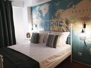 Bedroom with a white metal bed, green and white bedding, and a world map mural on the wall, lit by two hanging lamps.