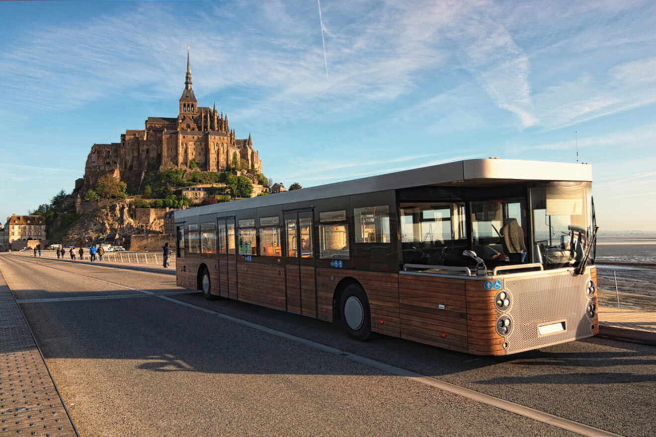 A bus is parked on a road near a large, historic castle-like structure built on a rocky island called Mont Saint Michel