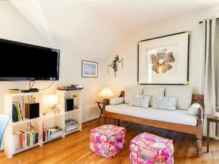 A cozy living room with a wall-mounted TV, a wooden couch with cushions, colorful ottomans, a bookshelf with books and decor, and framed artwork on the walls.