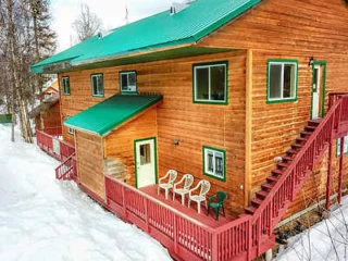 A wooden house with a green roof and a red deck is surrounded by snow. Four chairs are placed on the deck near the entrance. A staircase leads to the upper level.