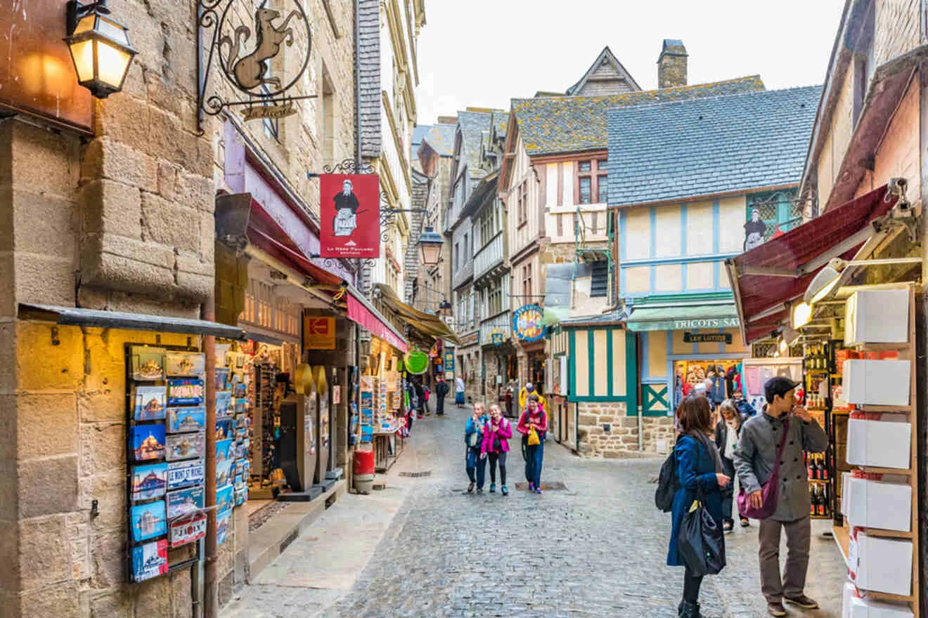 A cobblestone street lined with historic buildings, shops, and signs. People walk and browse, with a stone lantern on the left.