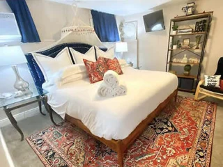 Cozy bedroom with a neatly made bed, patterned rug, wall-mounted TV, and shelving unit. Decor includes blue curtains, elegant lighting, and a heart-shaped towel on the bed.