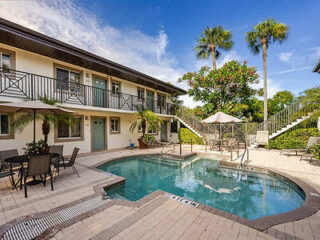 A small outdoor swimming pool surrounded by a patio area with tables and chairs, located in front of a two-story building with palm trees in the background.