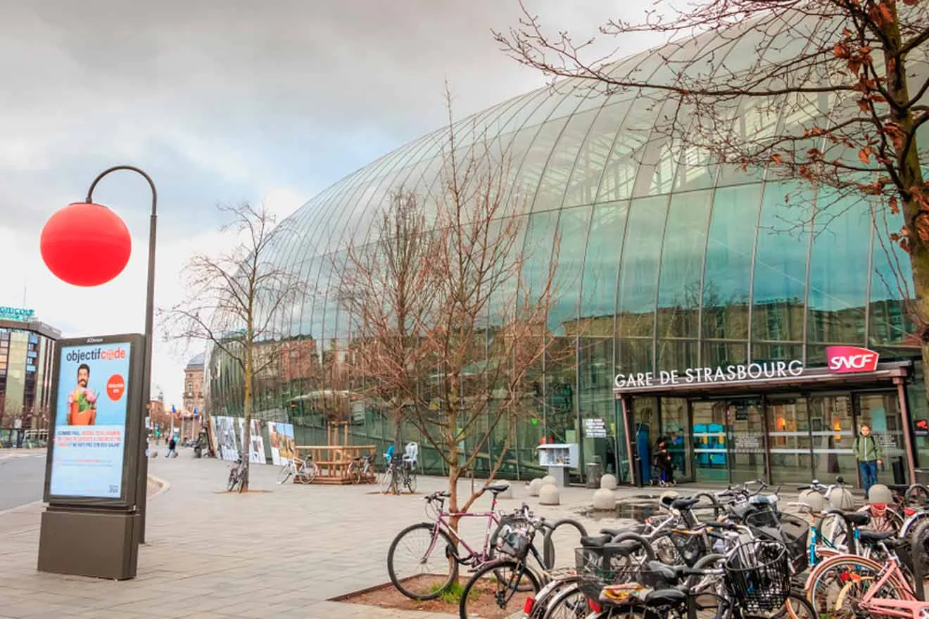 Modern glass entrance of Gare de Strasbourg with bicycles parked in front and a bright red streetlight globe on the left.