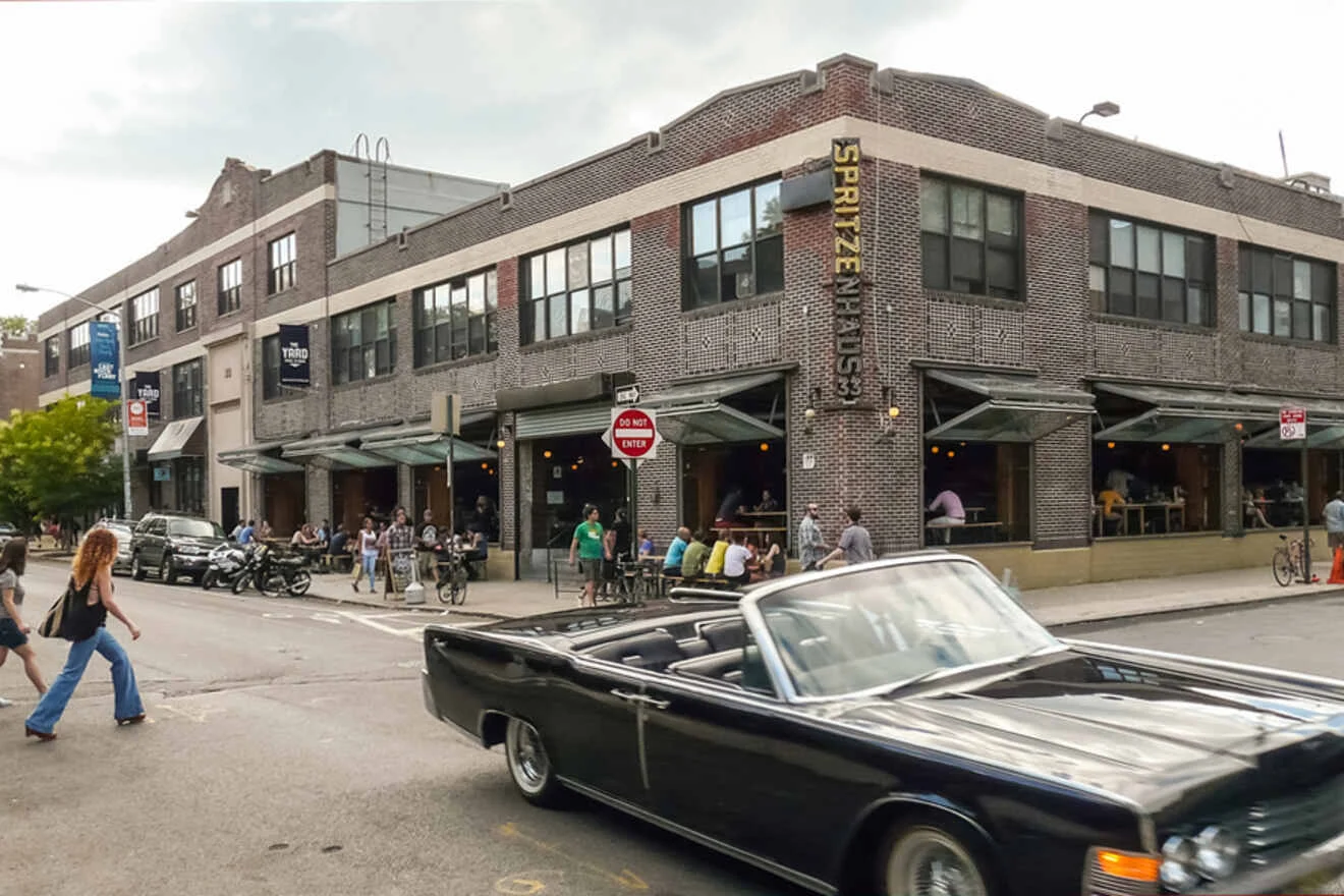 A classic black convertible drives past a busy corner with a brick building housing