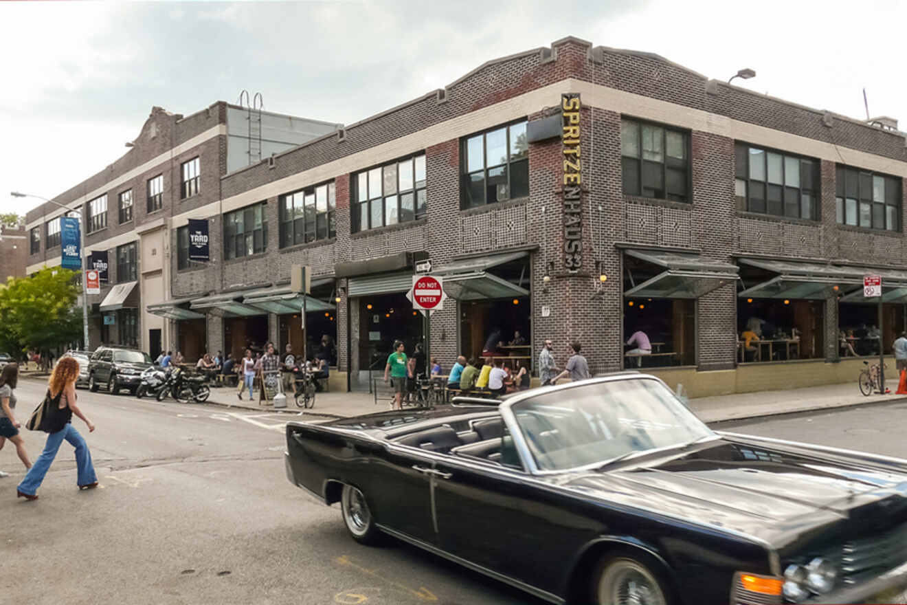 A classic black convertible drives past a busy corner with a brick building housing 