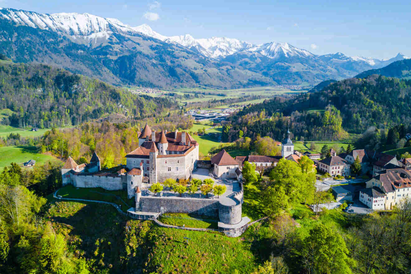 Aerial view of a historic castle surrounded by trees in Switzerland, mountains, and countryside with snow-capped peaks in the background under a clear blue sky.