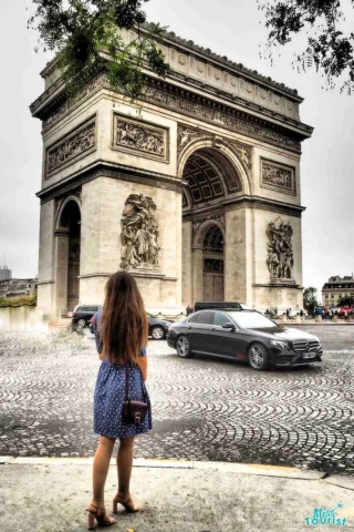 The writer of the post in a blue dress stands on a cobblestone street facing the Arc de Triomphe in Paris, with cars passing by.