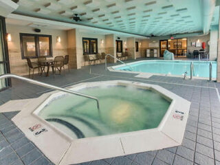 Indoor pool area with hot tub in the foreground and a swimming pool in the background. Tables and chairs line the wall, with ceiling lights providing illumination.