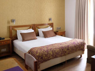 A hotel room with a double bed, white sheets, brown pillows, and a textured blanket. Wall lamps are above the bed, and there are curtains near a window.