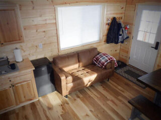 Cozy wooden interior with a small brown sofa, plaid pillow, closed window blinds, a mini fridge, and coats hanging by the door.