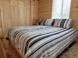 Bed in a wooden room with striped bedding, two pillows, a window with blinds, and a bedside lamp.