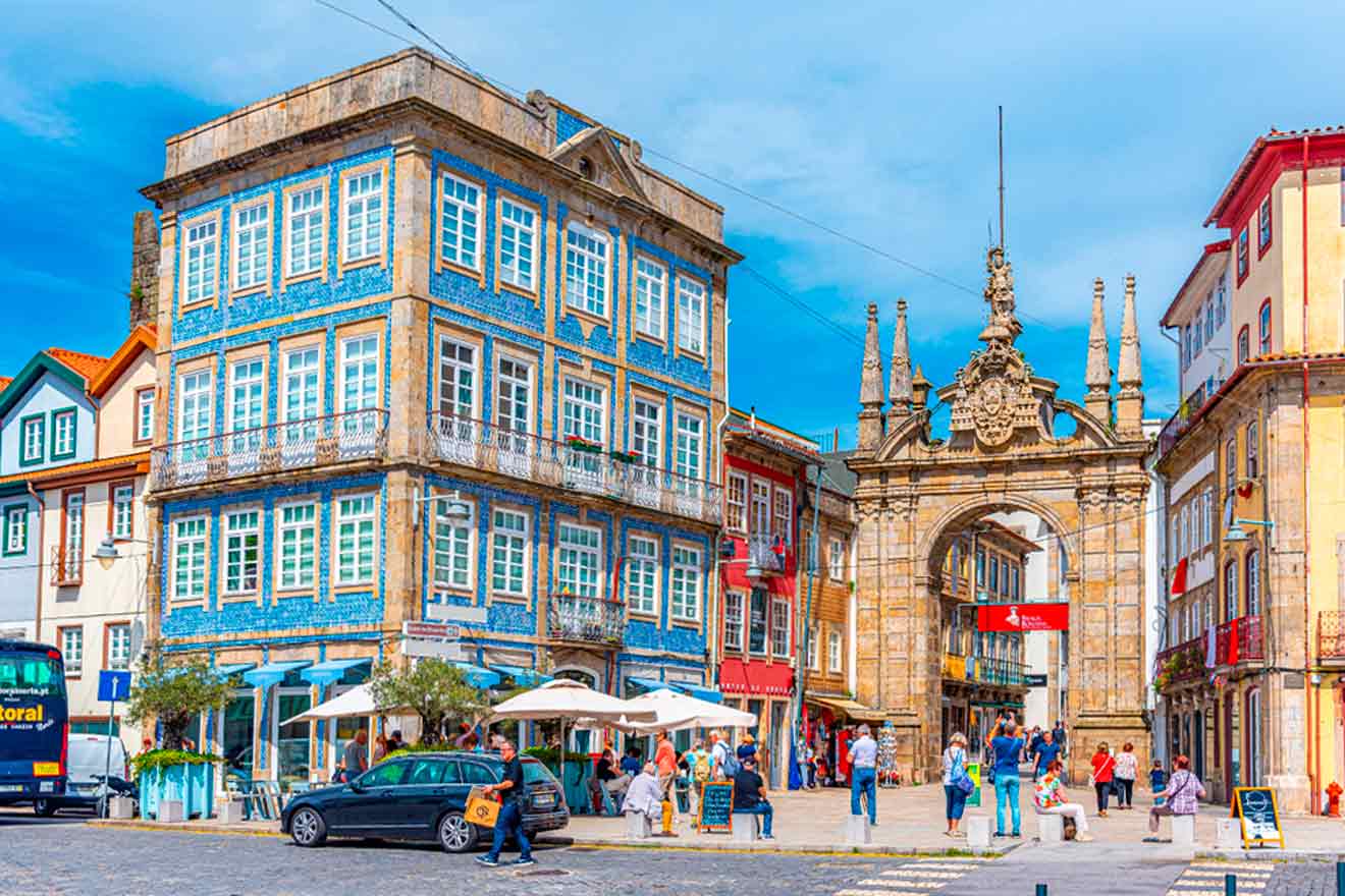 A historic European street scene with a blue-tiled building, a stone archway, and people walking and sitting at outdoor cafes.