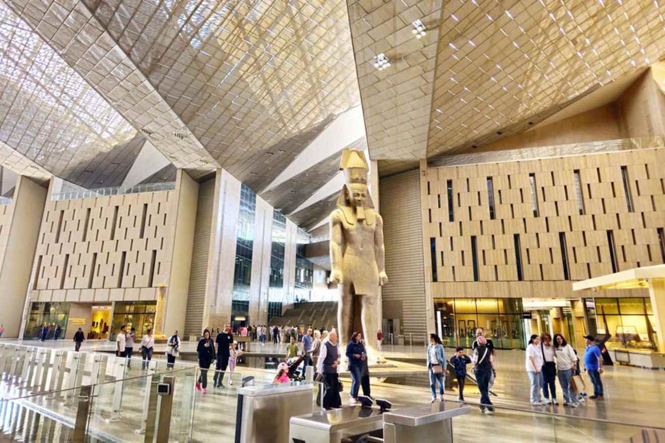 People walking inside a spacious, modern atrium with a large ancient statue at the center and geometric ceiling design above.