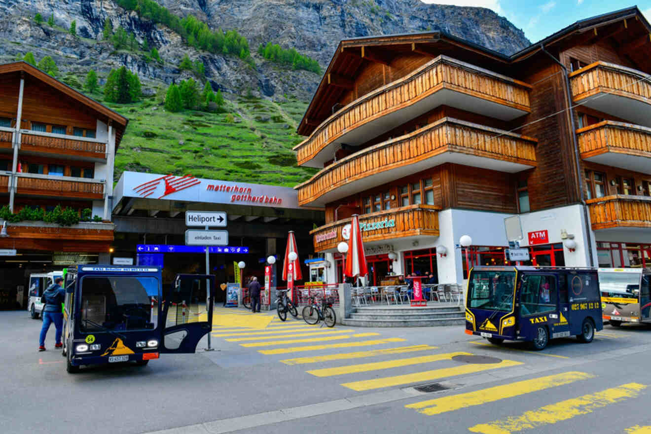 Street scene in Zermatt with electric shuttles, pedestrians, and wooden buildings. Signs for Matterhorn station, heliport, and an ATM are visible. Mountain backdrop included.