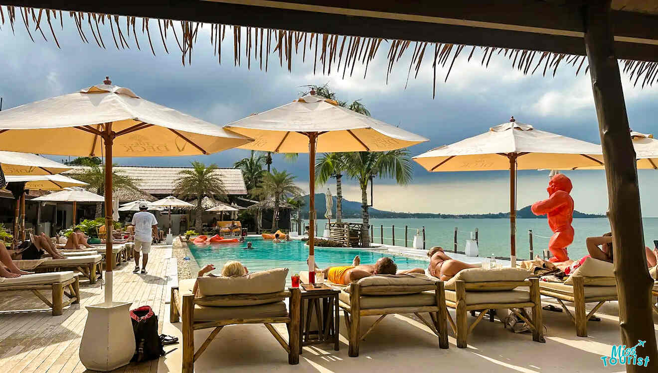 People lounging on poolside chairs under umbrellas with a view of the ocean in Koh-Samui. A large orange sculpture stands nearby under a cloudy sky.