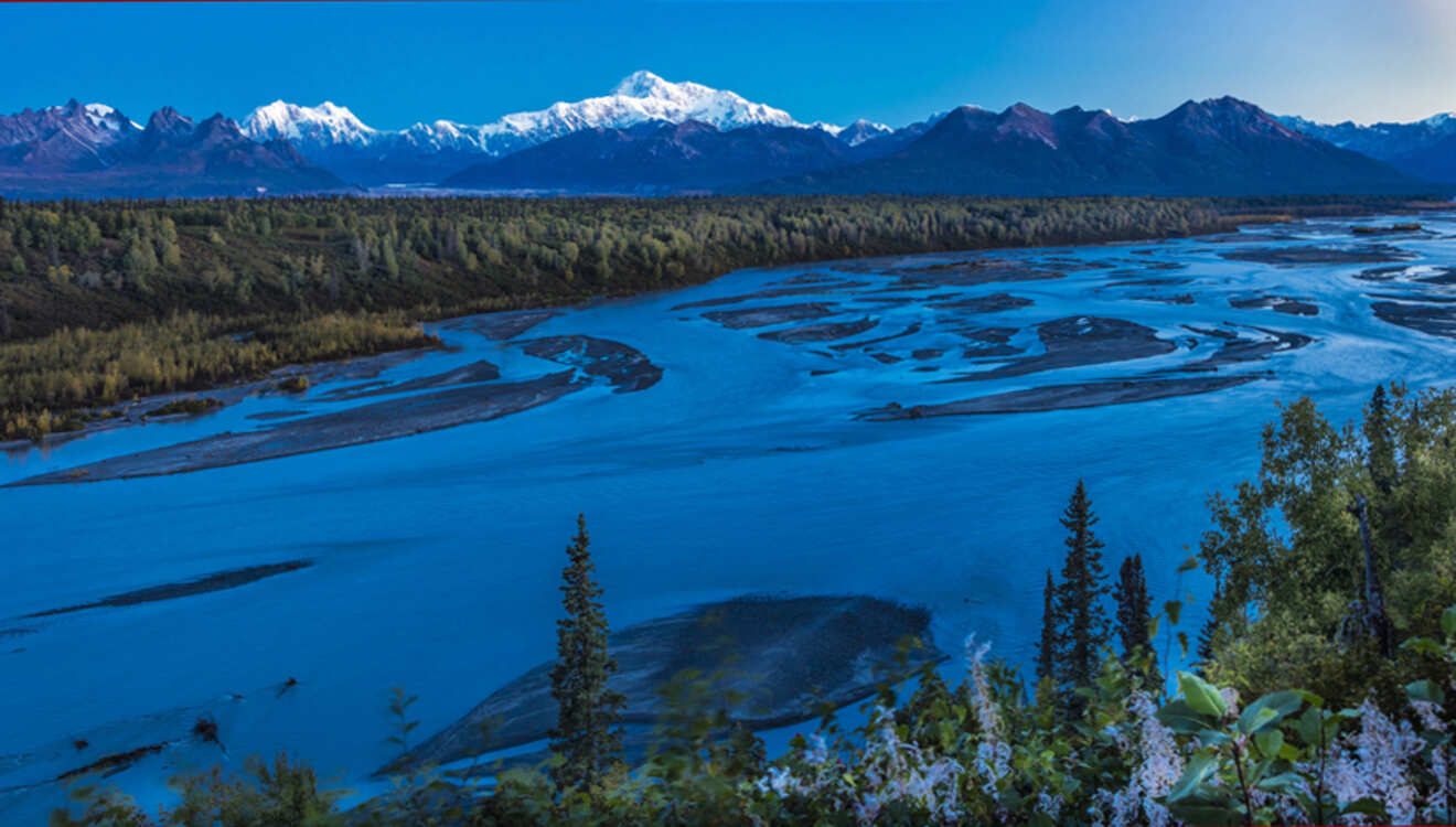 A vast blue river winds through a forested landscape with snow-capped mountains in the background under a clear sky.