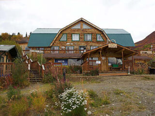 A rustic wood lodge with a green metal roof, surrounded by wildflowers and shrubs, set against a lightly clouded sky.