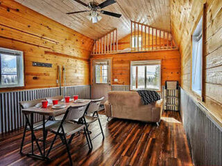 Wooden interior of a cabin with a dining table and four chairs, a sofa, ceiling fan, and a loft area. Natural light enters through the windows, illuminating the space.