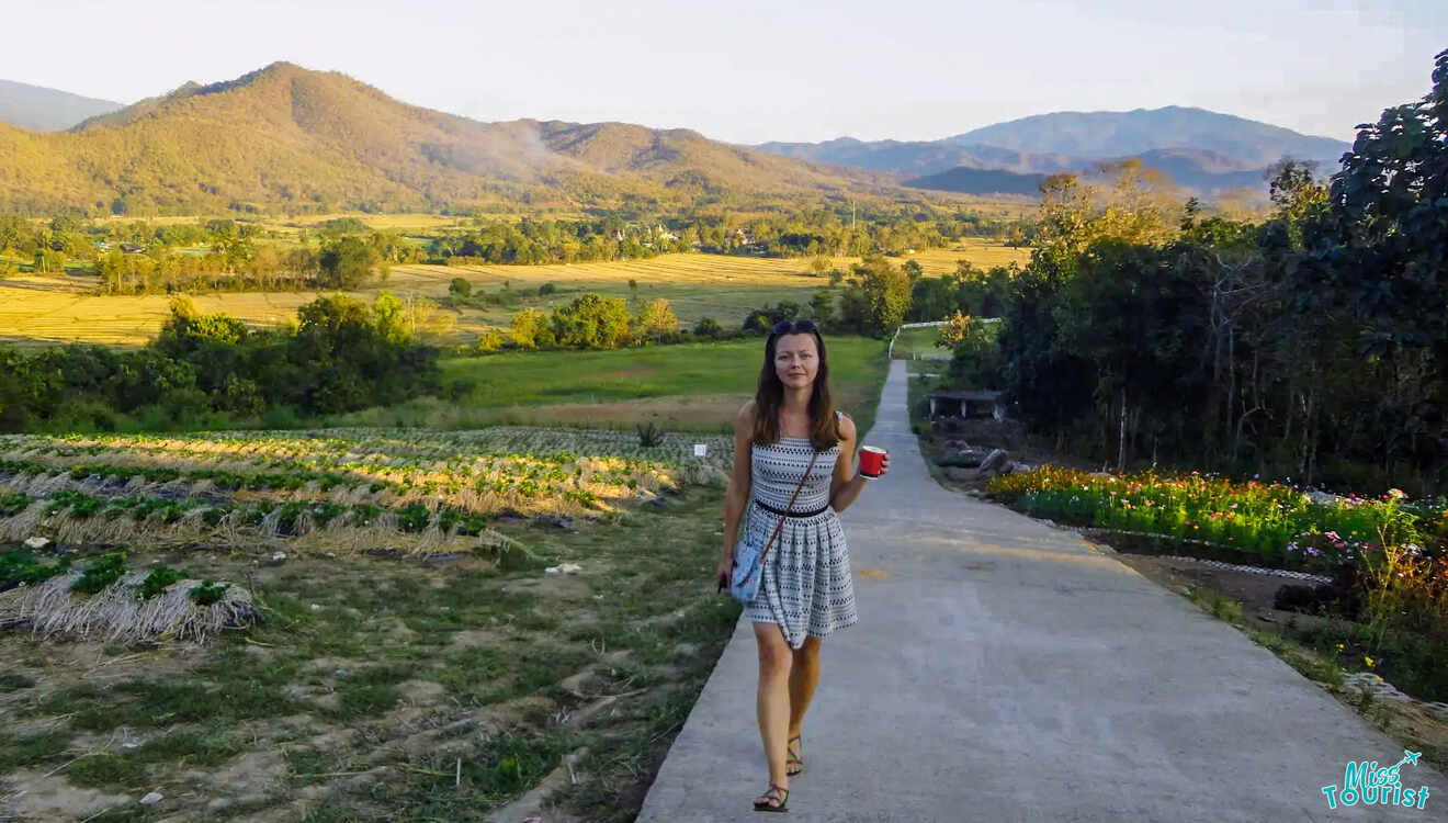 The writer of the post walks on a path through a scenic countryside, with fields, trees, and mountains in the background under a clear sky in Pai