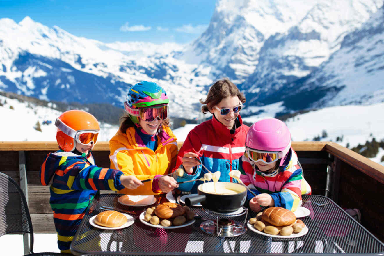 A family in colorful ski gear enjoys a fondue meal outdoors, surrounded by snowy mountains.