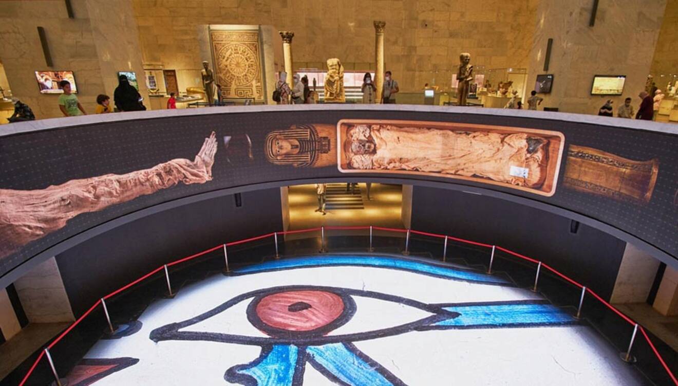 Museum interior featuring an exhibit with Egyptian artifacts. A digital display shows a mummy and sarcophagus, and the floor displays a large Eye of Horus design. Visitors are in the background.