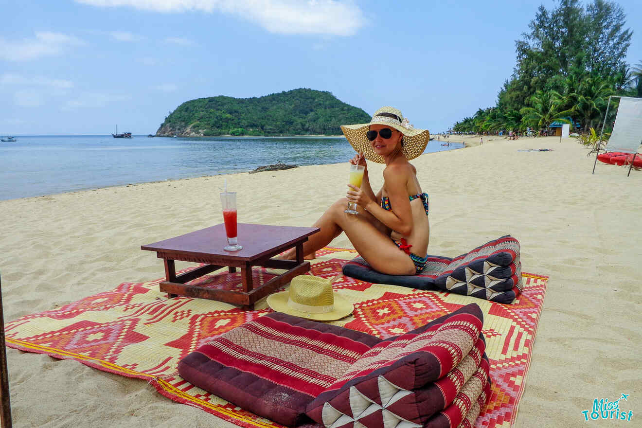 The writer of the post sitting on a beach at Koh-Phangan, wearing a hat and sunglasses, holding a drink. A patterned mat and cushions are on the sand nearby, with drinks on a low table. Sea and greenery in the background.