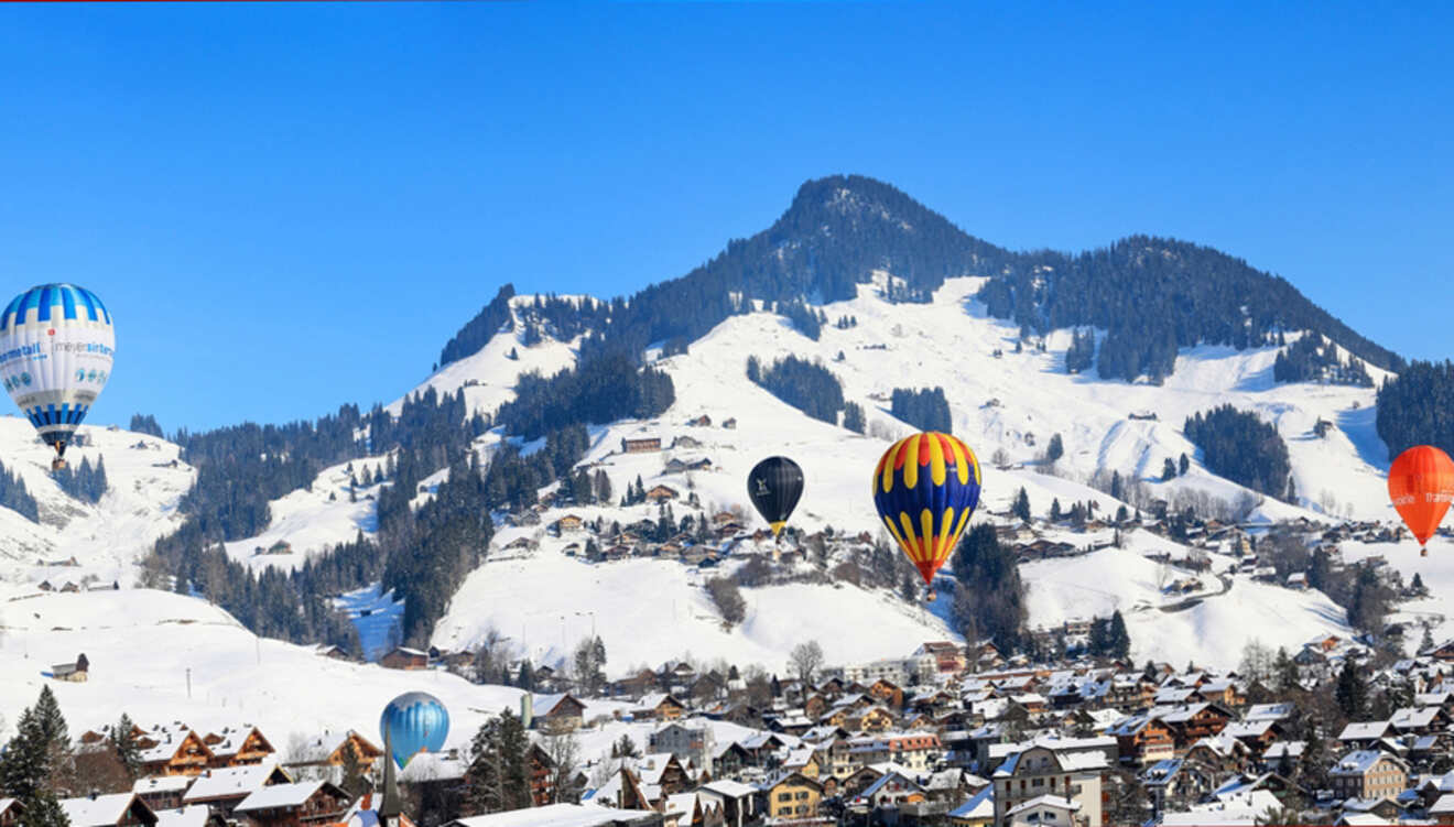 Hot air balloons float above a snowy mountain in Switzerland with scattered houses below under a clear blue sky.