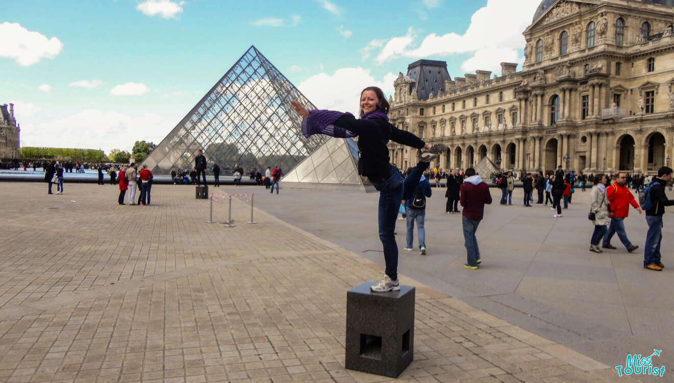 The writer of the post posing in a ballet stance on a pedestal near the Louvre Pyramid with people and historic buildings in the background.