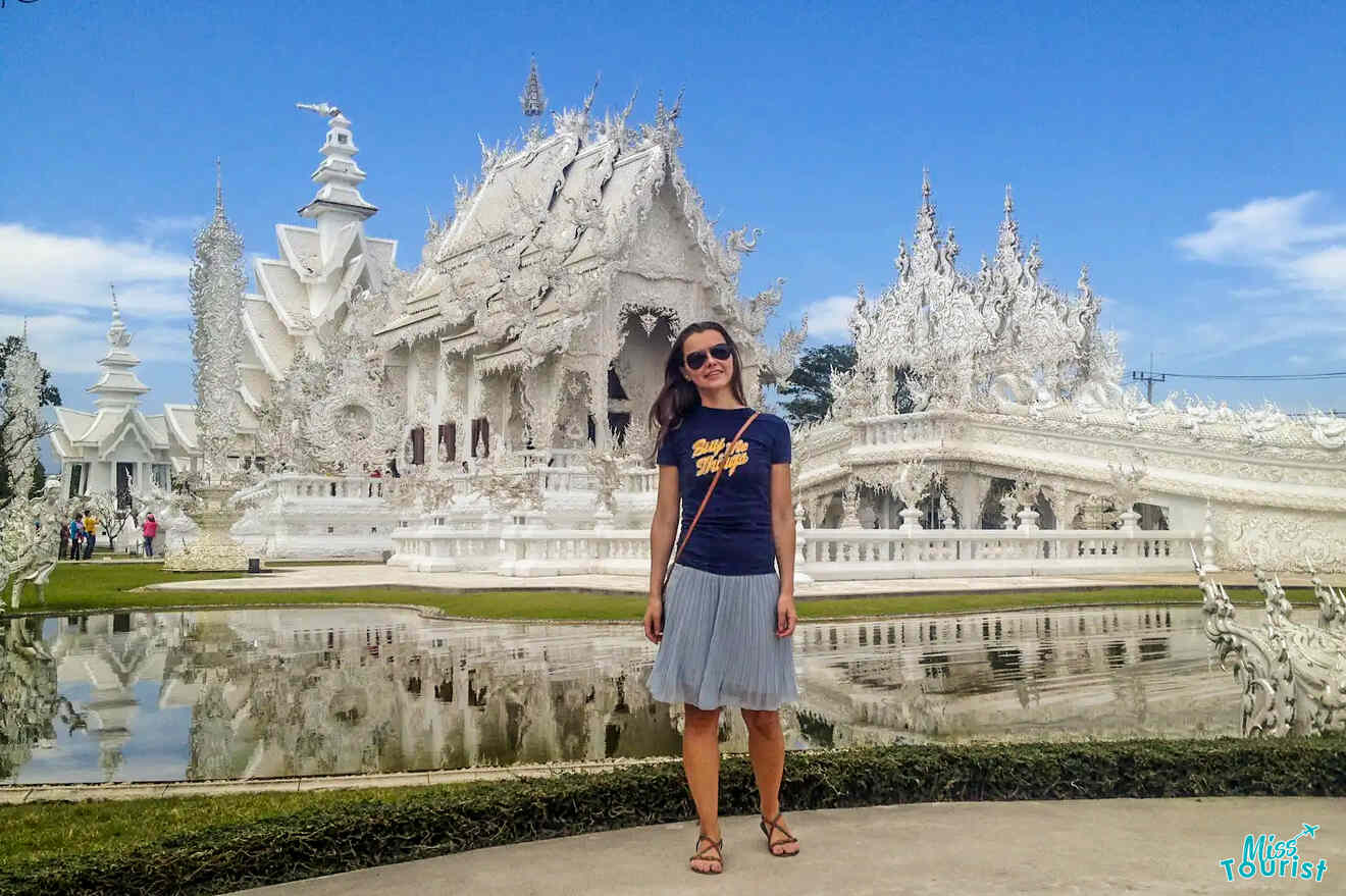 The writer of the post stands in front of an ornate Chiang-Rai-White-Temple-Wat-Rong-Khun with intricate designs and a reflecting pool on a sunny day.