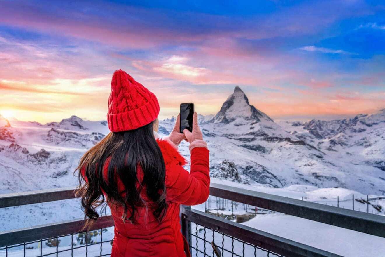 A person in a red coat and hat takes a photo of a snowy mountain in Switzerland, featuring a prominent peak, from a viewing platform.