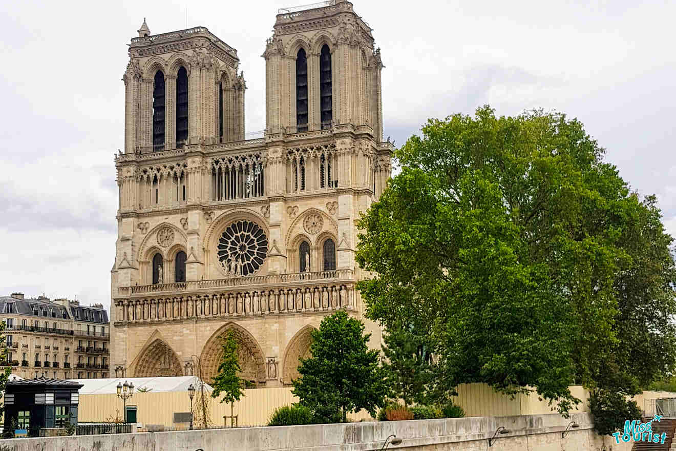 Notre-Dame Cathedral with its two towers is partially obscured by trees. The gothic facade and rose window are visible. Cloudy sky in the background.