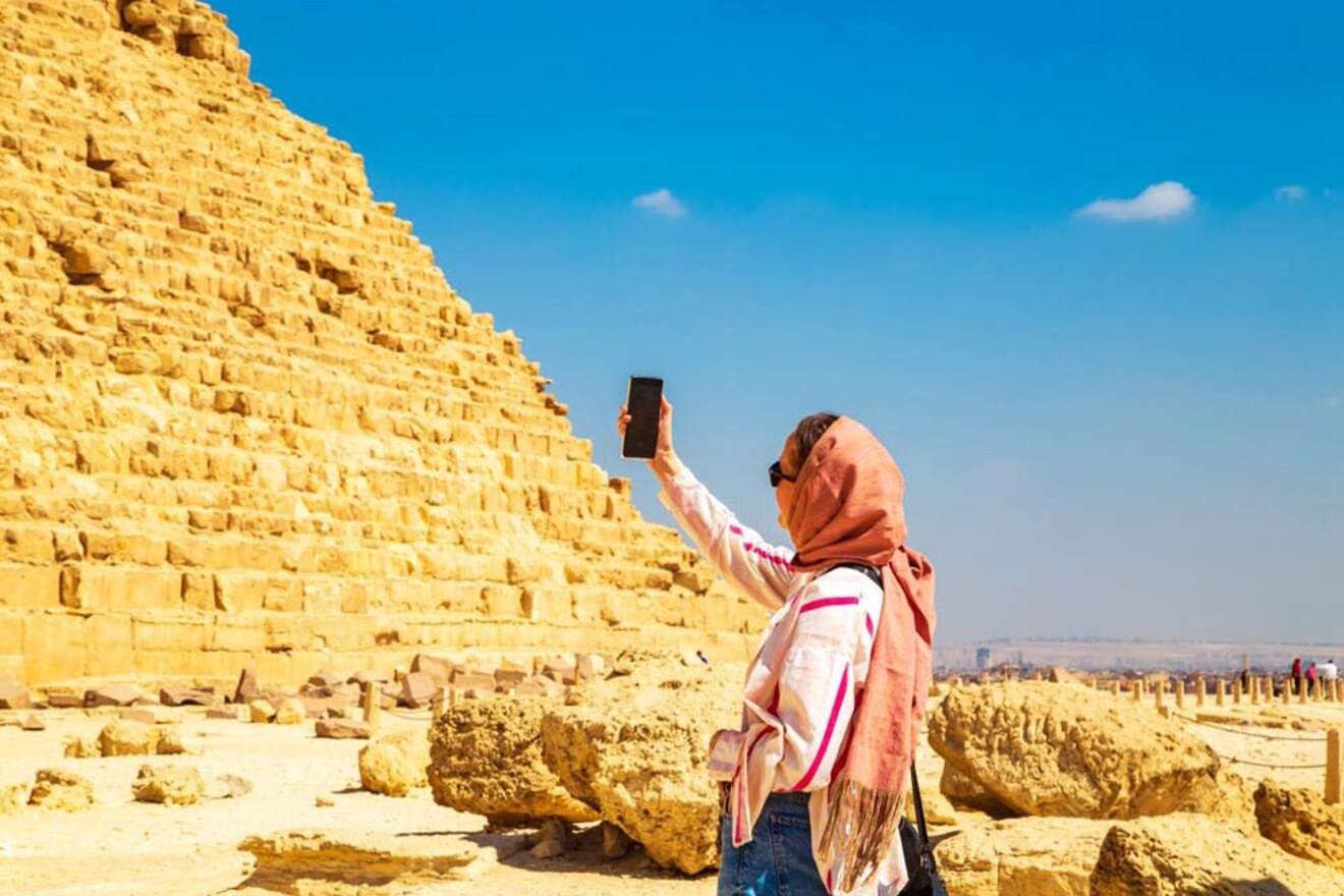 A person wearing a headscarf takes a selfie in front of an ancient pyramid under a clear blue sky.