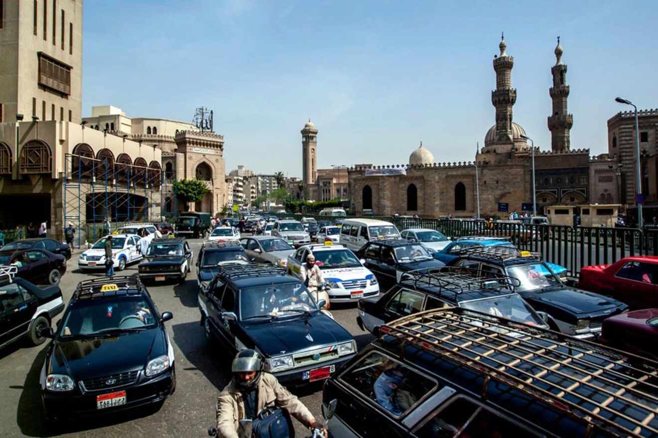 Busy city street with dense traffic, including cars and motorbikes. Historic stone buildings and mosques with minarets in the background under a clear blue sky.