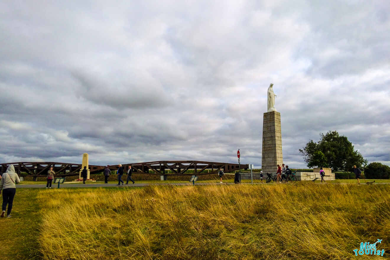 People walking near a tall statue on a grassy landscape with a cloudy sky in Normandy